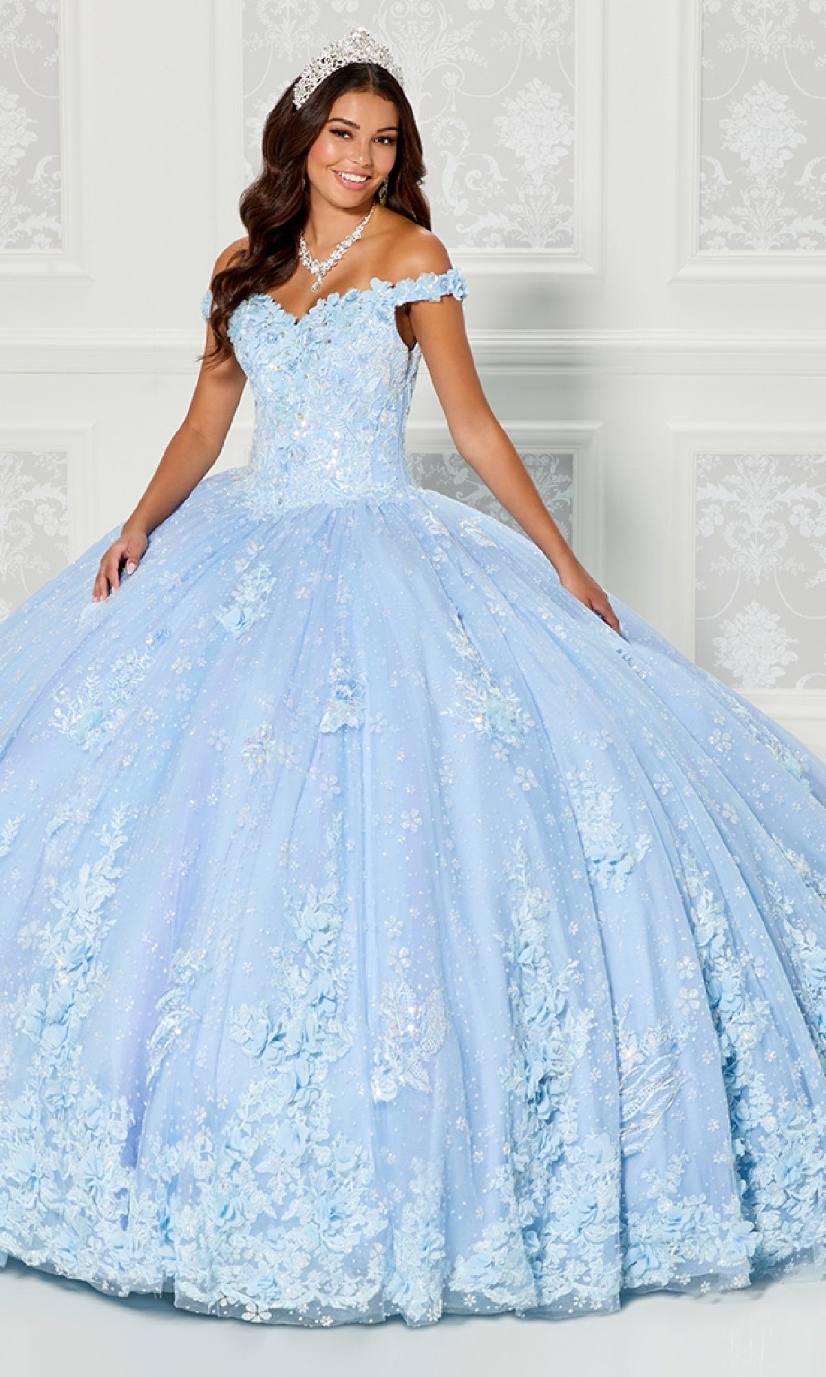 A woman stands indoors wearing the Quinceanera Dress PR30111 By Princesa, a light blue off-the-shoulder gown with floral appliqués and sequins, her long wavy hair adorned with a tiara.